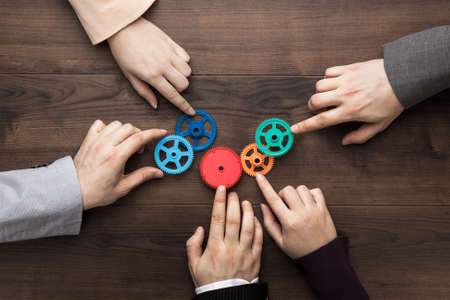 Teamwork concept. Different hands of men and women connect colorful gears into working mechanism on the brown wooden table background. Each has its own role in problem-solving. Experience exchange