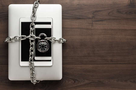 data security concept: computer, tablet, phone bound by metal chain and closed with combination lock on wooden tableの写真素材