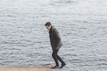 handsome young man in coat walking near waterの写真素材