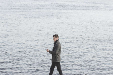 handsome young man in coat walking near waterの写真素材