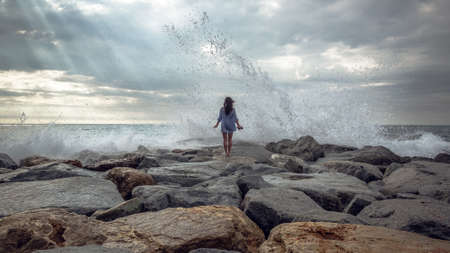 young woman standing on stones while big wave hitting shoreの写真素材