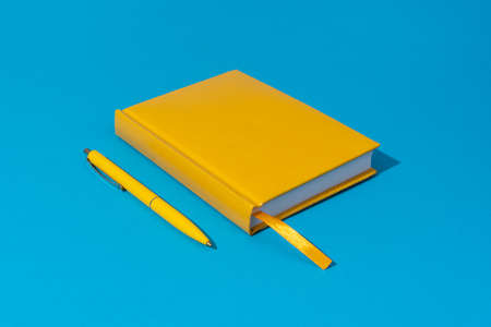 Photo of closed yellow notebook and ball-point pen over blue background. Minimalist image of closed diary and yellow pen as back to school concept.の写真素材