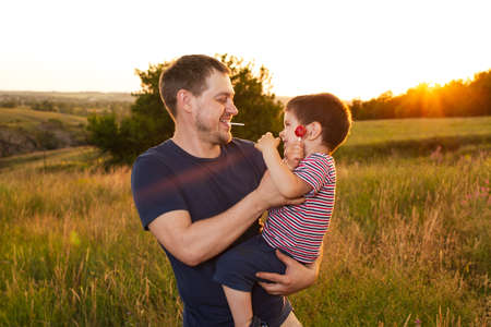 Dad and son walk in a yellow summer field in the evening at sunset and eat candy on a stick. Fathers Day, love in the family, the role of the father in raising the child.の写真素材
