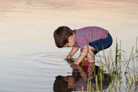 A little boy 2-3 years old plays on the river bank, the reflection of a child in the water. Outdoors in summer.の写真素材