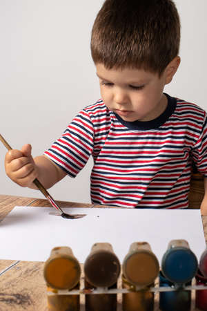A little boy 2-3 years old in a striped multi-colored T-shirt draws guasya paints on paper. Leisure...