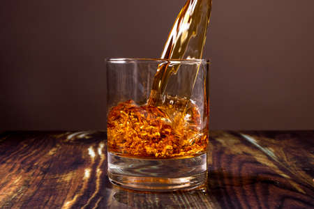 Whiskey is poured into a glass. Fiery high-end alcoholic drink in a bar on a dark wooden background.