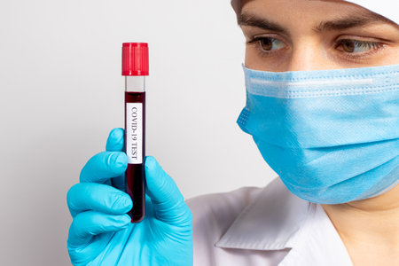 A doctor or lab technician wearing a mask and gloves is holding a blood test tube with the...
