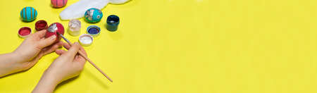 A woman paints a chicken egg with gouache paints with a brush on a yellow background. Easter photo, preparation for the holiday.の写真素材