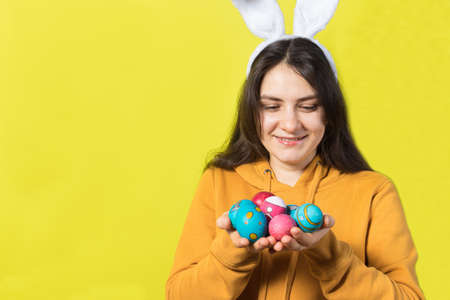 A happy brunette woman with rabbit ears on her head holds Easter eggs and smiles. Prepare for...