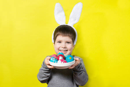 Cheerful happy boy with bunny ears on head is playing with Easter colorful eggs, beating painted chicken eggs against each other. Easter kid on a yellow backgroundの写真素材