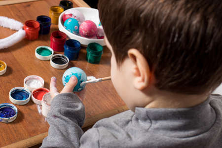 A little boy paints gouache eggs. Preparing for Easter, cheerful happy child painted with the colors of a chicken egg.の写真素材