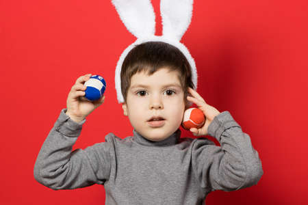 Cheerful happy boy with bunny ears on head is playing with Easter colorful eggs on a red background...