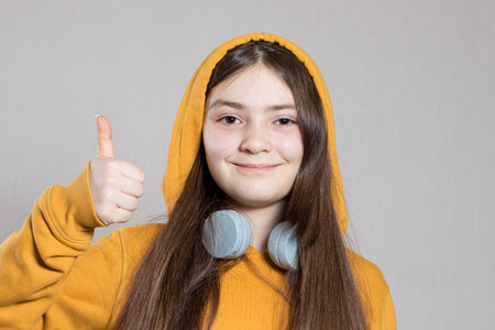 A teenage girl in a bright hooded sweatshirt on her head and headphones shows off her finger up the wow. Learning online, schooling remotely. On a light background, copy space for text.の写真素材