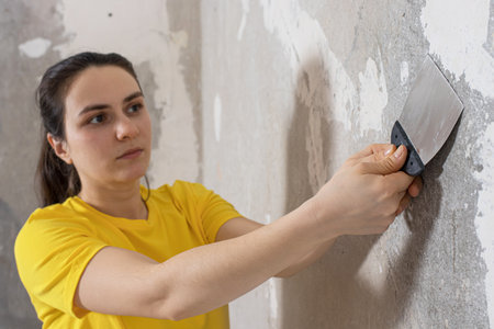 The repairman removes old dangling wallpaper from the wall with a spatula. Removing wallpaper residues during repairs.の写真素材