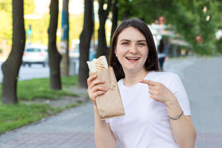 A beautiful brunette girl keeps a shawarma in the street. Food on the way on the way to work. Fast food cafe advertising.の写真素材