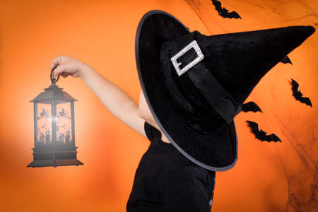 A little boy 4 years old in a witch costume in a black hat holds a glowing lantern on an orange background. Halloween kids.の写真素材
