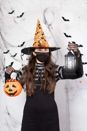Halloween masked people. A woman in a witch costume in a protective mask holds a pumpkin candy and a luminous lantern. During the pandemic vertical photoの写真素材