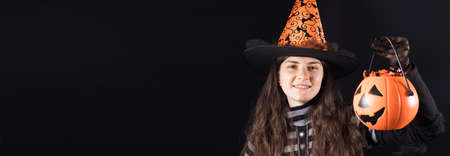 Halloween people banner. A woman in a witch costume on a black background holds bucket pumpkin with candy and smile. Place for copy space text.の写真素材