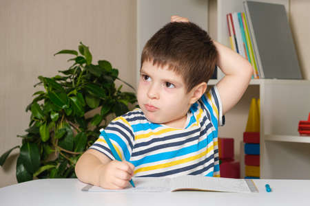 A boy in a striped T-shirt scratches his head, writes spells, prepares for school.