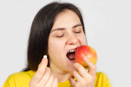 A woman bites a red apple while holding a multivitamin pill in her handの写真素材