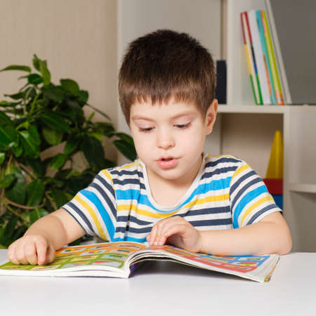 A 4-year-old child sits at a table and reads a book, looks at pictures, learns to read.の写真素材