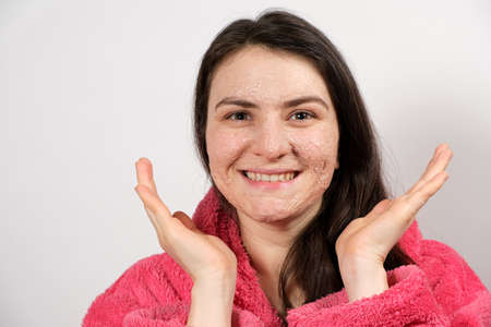 A young brunette woman in a pink bathrobe makes cleansing facial treatments - a mask or scrub to cleanse and nourish the skin.の写真素材