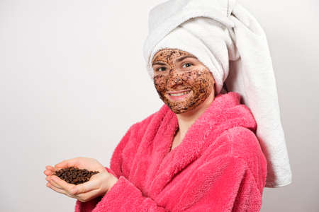 A woman in a pink bathrobe with a coffee mask on her face holds coffee beans in her handsの写真素材