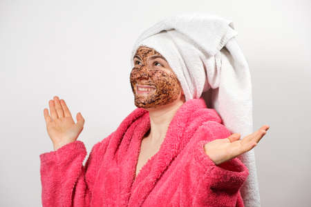 A beautiful woman with a coffee scrub on her face in a bathrobe with a towel on her head on a white background makes spa skin care proceduresの写真素材