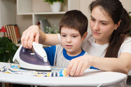 A mother teaches her 4-year-old son to iron clothes. Helping mom with the housework, a little helperの写真素材