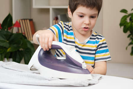 A boy of 4 years learns to hold an iron, iron clothes on an ironing board. Helper, help children to care for the house.の写真素材