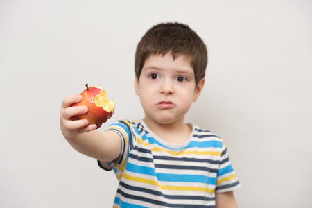 A 4 year old boy eats a red apple and squirms. The child does not like fruitの写真素材