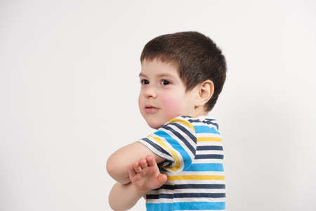 A happy boy of 4 years old in a striped T-shirt holds his hands near his chest and looks sideways, a...