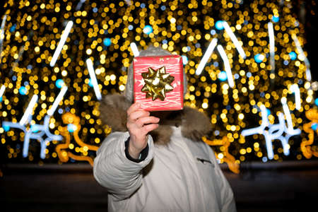 A woman holds a red box with a gift instead of a head standing near a Christmas tree with a yellow...