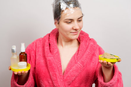 A woman with a film on her head makes a hair mask and holds natural and unnatural cosmetics in her handsの写真素材