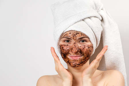 A young woman with a coffee mask on her face and a bath towel on her head smiles and looks into the camera on a white background, a place for text.の写真素材