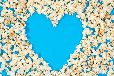 Popcorn is laid out in the form of a heart with a place for text on a blue background, top view.の写真素材