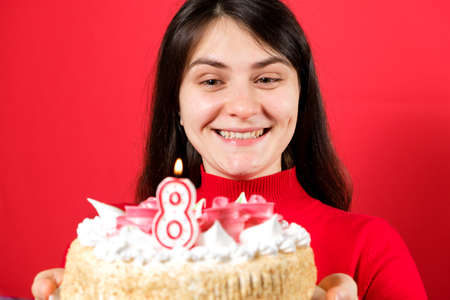 A happy woman holds a cake with a candle in the shape of the number eight on a red background