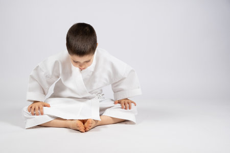 A little boy in a kimono practices karate, sits in a butterfly pose and practices stretching, a place to textの写真素材