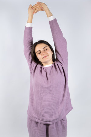 Young brunette woman in muslin lavender pajamas sleep clothes stretches by raising her arms up and closing her eyes.の写真素材