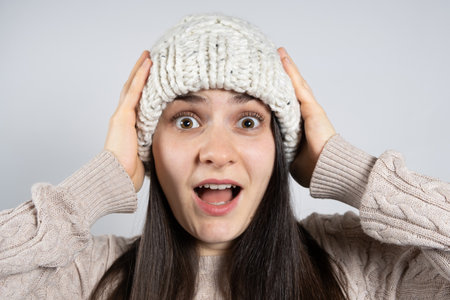 A young brunette woman in a hat and sweater looks into the camera and screams holding her hands to her head.の写真素材
