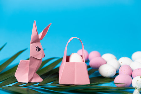 An Easter bunny figurine made of paper, origami, and a basket with eggs on a blue background, place...