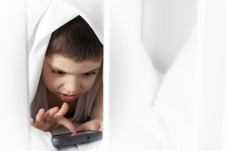 A 5 year old boy plays games on his phone while lying in bed. Gadgets in bed before bedtime in a childの写真素材