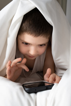A 5 year old boy plays games on his phone while lying in bed. Gadgets in bed before bedtime in a child.の写真素材