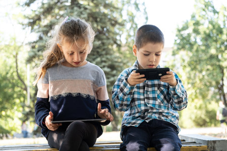 Children use gadgets while sitting in the park outdoors, games on smartphone and tabletの写真素材