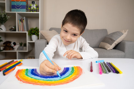 A small child draws a rainbow with felt-tip pens in a sketchbook while sitting at a tableの写真素材