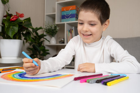 A small child draws a rainbow with felt-tip pens in a sketchbook while sitting at a tableの写真素材
