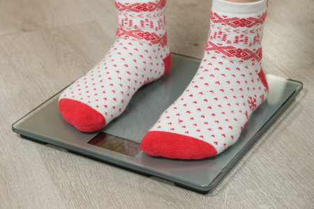 Feet in Cozy Christmas Socks Standing on Digital Bathroom Scales, New Year's Resolution Concept