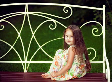 Beautiful little girl with long hair in the summer sitting on a bench and looks straight at the camera, mysteriously smilingの写真素材