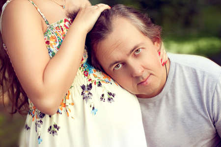 Husband middle age listening to stomach of pregnant wife and looking directly at the camera, touching, smiling, during a family vacation in the Park. Floral sundressの写真素材