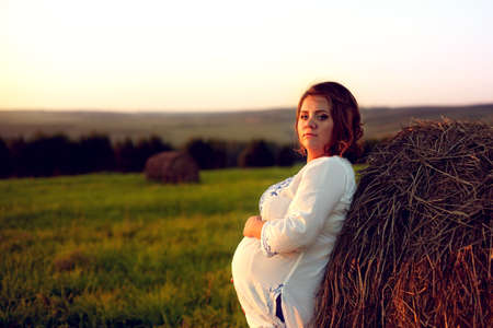 Young Pregnant Woman Standing by the Haystack at Sunset and Embracing her Belly. 8 Month Pregnancy. Happy Maternity Conceptの写真素材